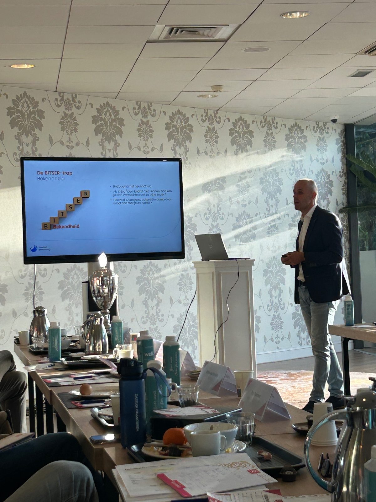 Man presenteert tijdens pitch van ProjectMaat bij BNI Ede met visuele ondersteuning op groot scherm en notities op tafel.